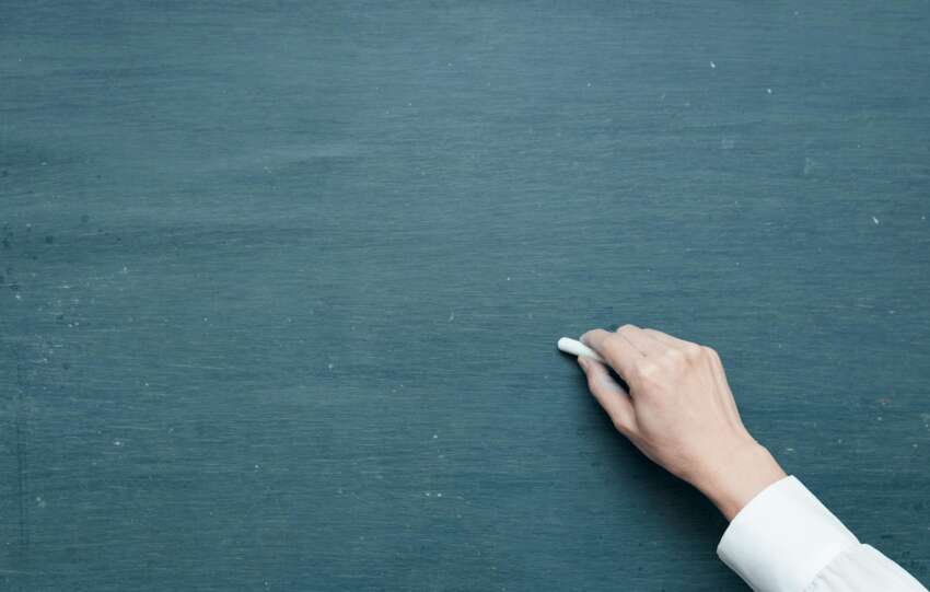 Hand holding chalk ready to write on a blank blackboard surface.