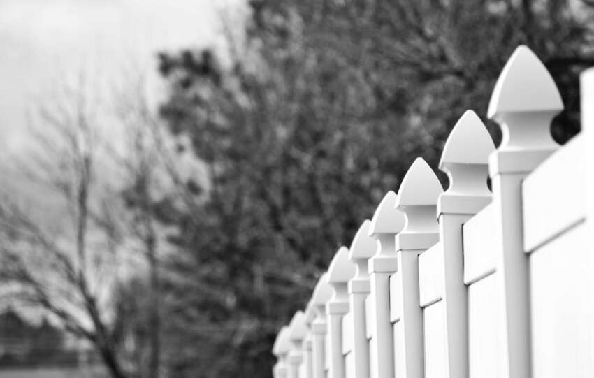 Black and white image of a row of decorative fence posts with blurred winter trees in the background.