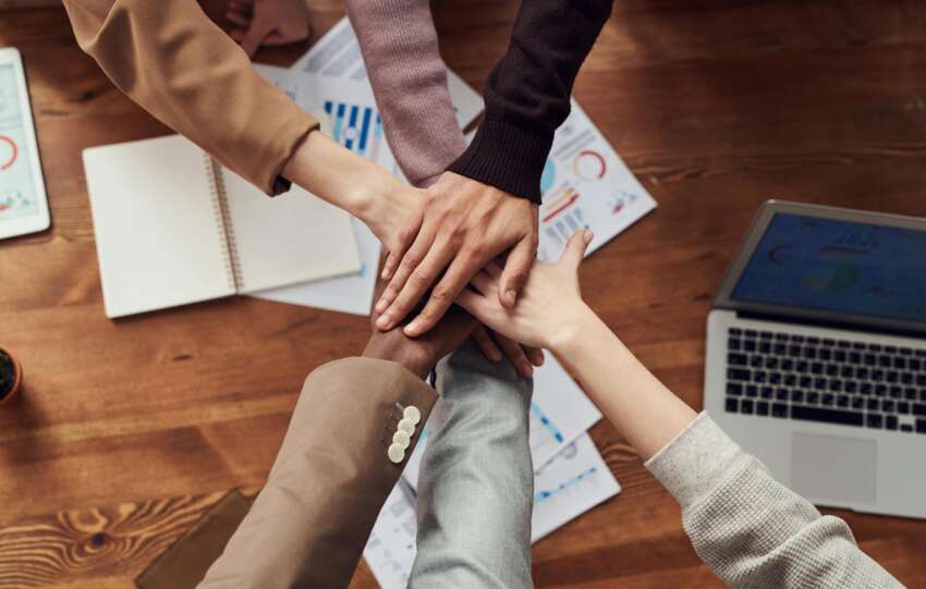 Diverse professionals unite for teamwork around a wooden table with laptops and documents.