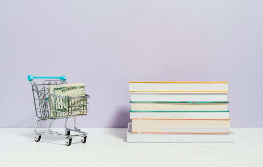 A shopping cart with dollar bills next to a stack of textbooks on a light background.