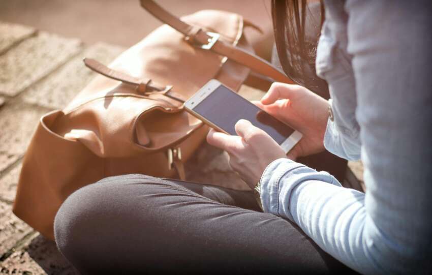 Woman sitting with smartphone and brown bag in sunlight.