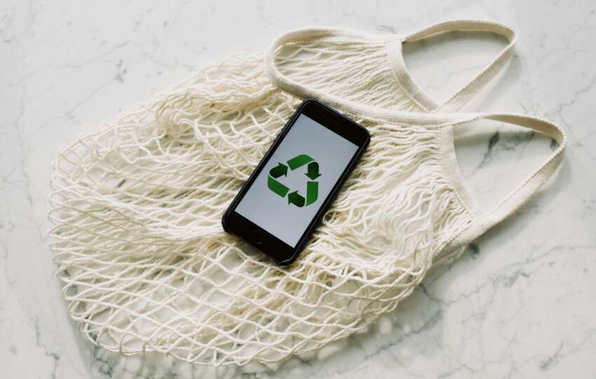 Overhead of smartphone with simple recycling sign on screen placed on white eco friendly mesh bag on marble table in room