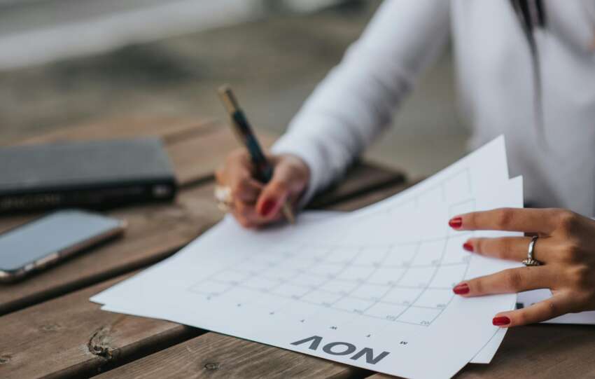 A woman planning with a calendar outdoors, jotting down notes on a wooden table with a pen.