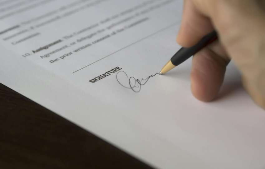 A hand signs a formal contract with a pen on a wooden desk.