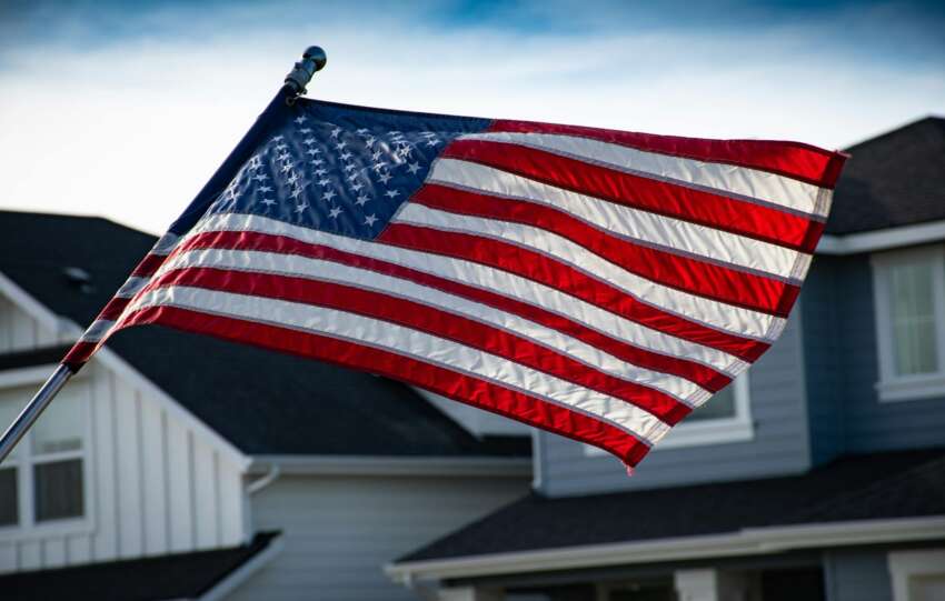 A close-up of the American flag waving in front of a suburban house, symbolizing patriotism.