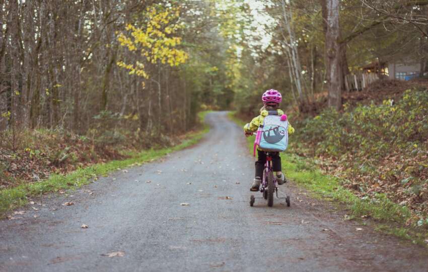 A young girl riding a bike with training wheels on a rural path surrounded by fall foliage.