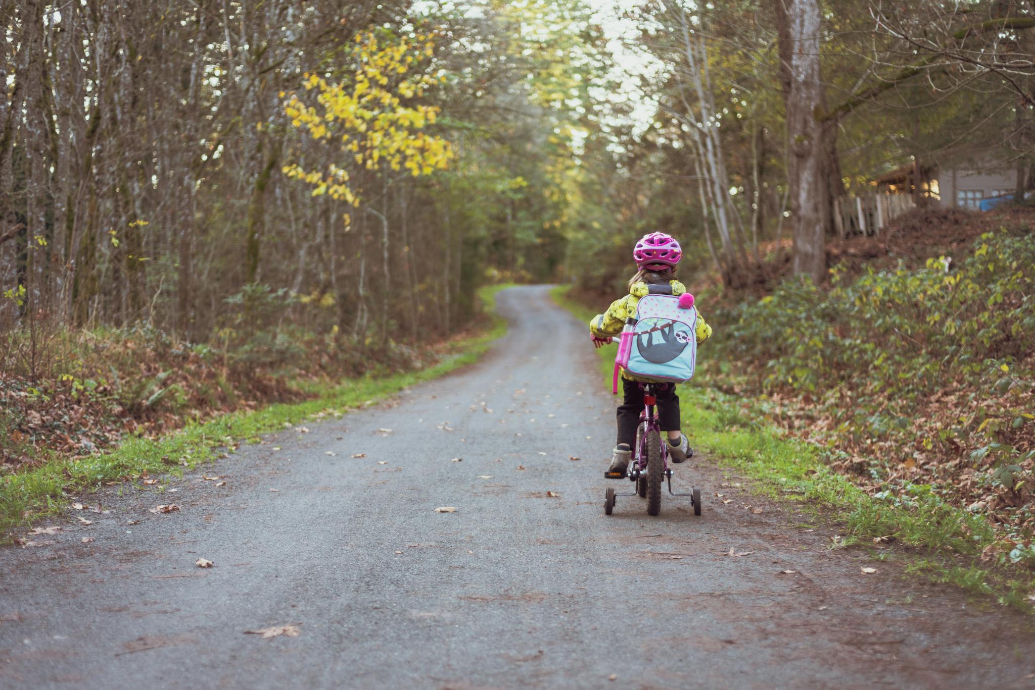 A young girl riding a bike with training wheels on a rural path surrounded by fall foliage.