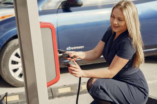 Side view of positive female in casual dress sitting near station on street charging electric vehicle for continuation trip