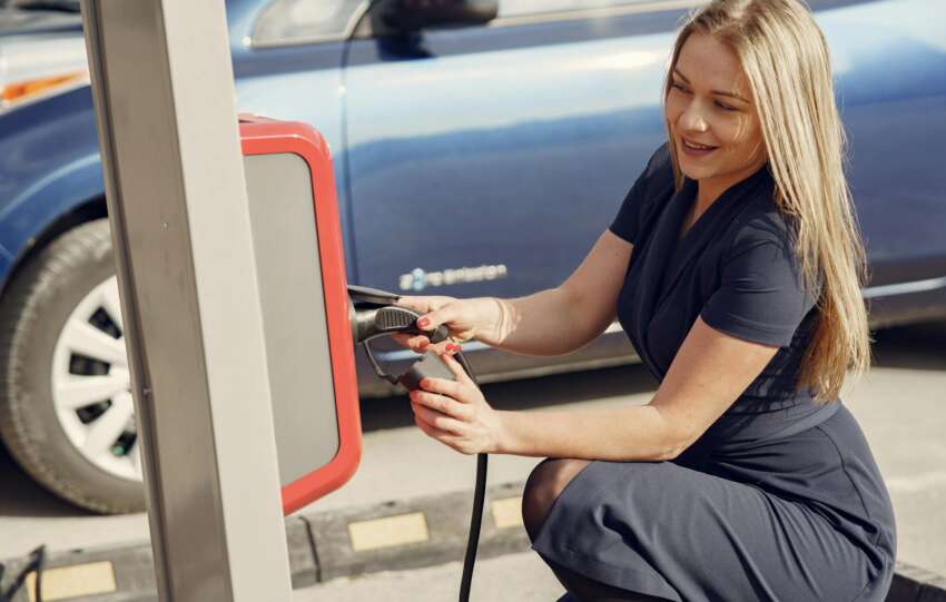 Side view of positive female in casual dress sitting near station on street charging electric vehicle for continuation trip