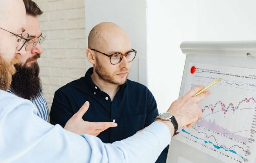 Three men discussing financial charts on a whiteboard during a business meeting.